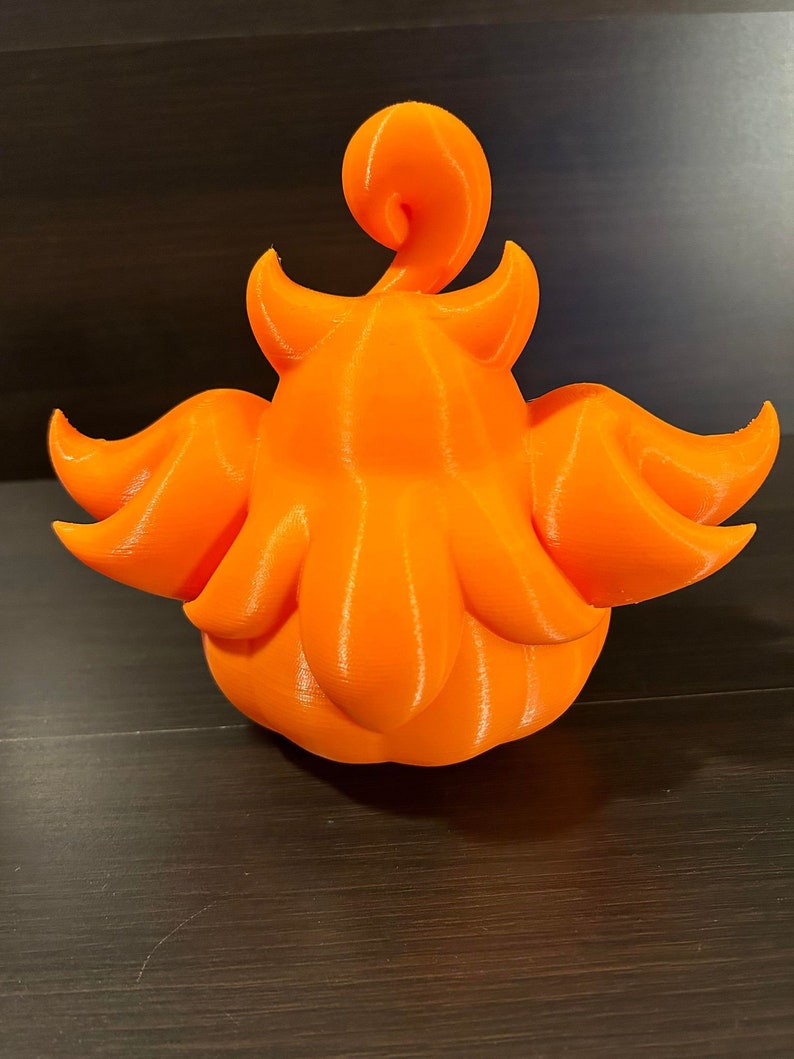 May include: A 3D-printed orange decorative object with a unique, stylized design. The object features a rounded base with petal-like structures and a curled top element. The bright orange color and smooth texture are prominent.