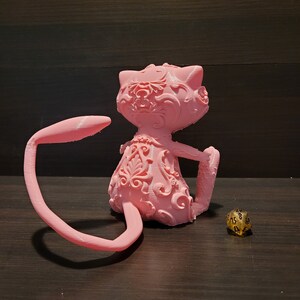 Op de afbeelding: Een roze 3D-geprint beeldje van een kat met ingewikkelde bloemmotieven. De kat zit met zijn staart om zijn lichaam gekruld. Een gouden 20-zijdige dobbelsteen bevindt zich op de voorgrond.
