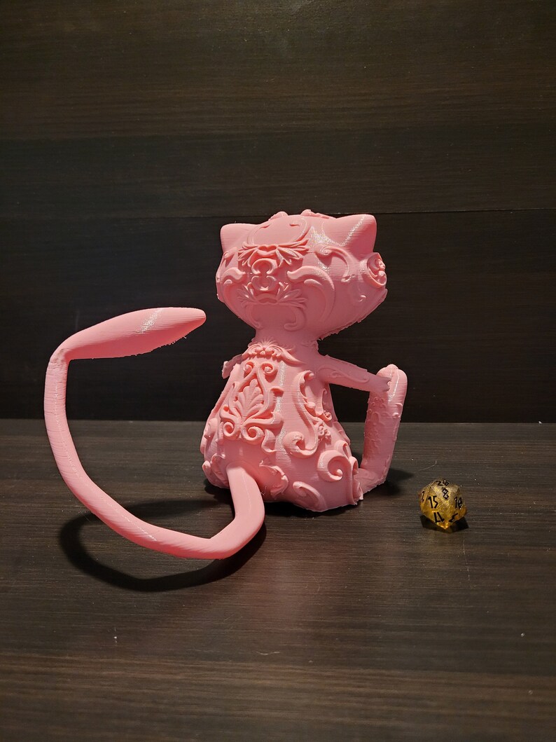 Op de afbeelding: Een roze 3D-geprint beeldje van een kat met ingewikkelde bloemmotieven. De kat zit met zijn staart om zijn lichaam gekruld. Een gouden 20-zijdige dobbelsteen bevindt zich op de voorgrond.