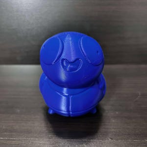 May include: A blue 3D printed figurine of a cartoon character with large eyes and a wide smile. The character has a round body and short legs.