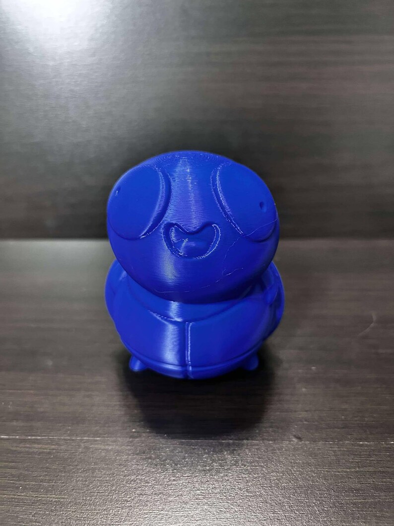 May include: A blue 3D printed figurine of a cartoon character with large eyes and a wide smile. The character has a round body and short legs.