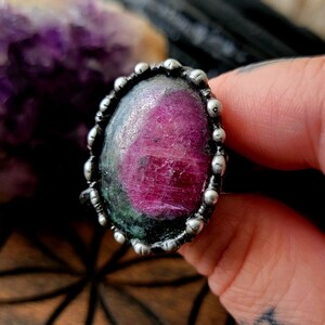 May include: A silver ring featuring a large, oval-shaped gemstone with a vibrant pink and green color. The gemstone is set in a textured silver band with small, round embellishments. The ring is held up by a hand.