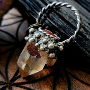 May include: A handcrafted pendant featuring a clear quartz crystal with a silver-toned metal setting. The setting has a textured design with small, round embellishments and a twisted loop for hanging. The crystal has a warm, amber hue.