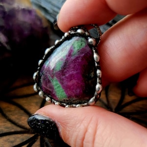 May include: A triangular pendant with a vibrant ruby gemstone, showcasing deep purple and green hues. The stone is set in a silver-toned bezel with a beaded edge. The pendant is held by a hand with black nail polish. The text "@crookedwitch fingers" is visible.