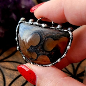 May include: A pendant with a silver-colored metal frame. The pendant features a polished, semi-circular stone with brown and black patterns. The stone has a decorative design in the center. The text "@crooked.witch.fingers" is visible.