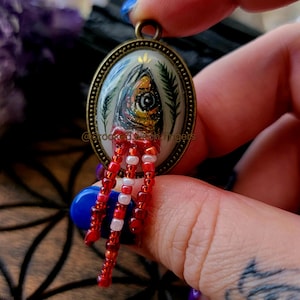 May include: Oval pendant with a painted fish design in a brass-colored setting. The fish has a yellow eye and red beaded fringe. The background is a mix of green, blue, and white. The pendant is held by a person.