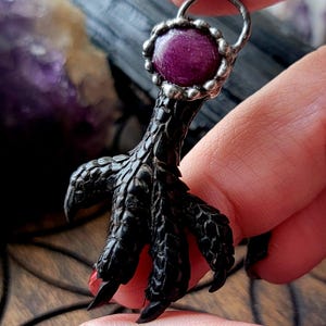 May include: A silver pendant featuring a black claw design with a round, purple gemstone. The claw has detailed texture and is held by a silver loop. The text "@crooked.witch.fingers" is visible.