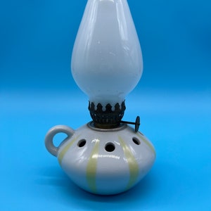 Op de afbeelding: Een witte keramische olielamp met een geel gestreept ontwerp en een glazen schoorsteen. De lamp heeft een handvat en een metalen lont houder.