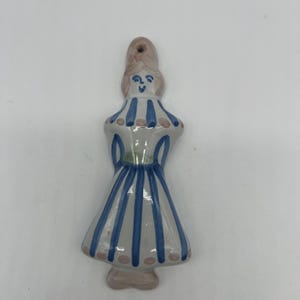 Puede incluir: Figura de cerámica de una mujer con un vestido a rayas azules y blancas. La figura tiene un agujero en la parte superior para colgarla. El vestido tiene detalles en rosa y un cinturón verde. El pelo de la mujer es castaño claro.
