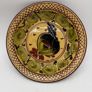 Puede incluir: Un plato redondo de cerámica pintado a mano con un diseño de gallo. El plato presenta un centro amarillo con un gallo negro y detalles rojos. El borde tiene un patrón de celosía marrón y espirales verdes y marrones.