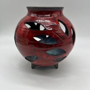May include: A red ceramic vase with leaf-shaped cutouts. The vase has a round body and a dark rim, supported by three small feet. The glossy finish has a marbled effect, with dark red and black tones.