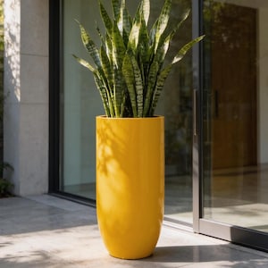 May include: A tall, cylindrical, glossy yellow planter containing a snake plant with upright, variegated green and yellow sword-shaped leaves. The plant is positioned on a light-colored stone surface in front of a large glass sliding door.