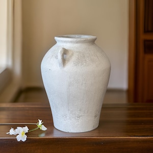 May include: A white ceramic vase with a textured finish and a handle. The vase is sitting on a wooden table.