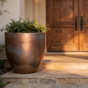 May include: Two large, rounded, metallic copper-toned planters with a weathered patina finish, each containing a lush green shrub, positioned on either side of a set of double wooden doors set within a stone archway of a building entrance.