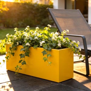 May include: A rectangular yellow planter box filled with green ivy and small white flowers. The planter has a smooth, solid-colored finish and sits on a dark stone patio surface, positioned next to a dark gray outdoor lounge chair.