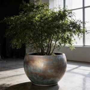 May include: A large, spherical planter with a weathered, metallic finish featuring blended tones of bronze, copper, and oxidized teal green. The planter holds a dense, leafy green shrub with thin, branching stems that fill the upper portion.