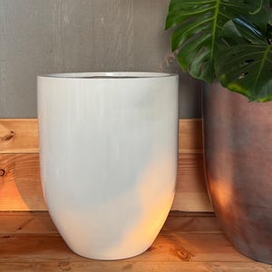 May include: A tall, white, glossy ceramic planter with a rounded base and a slightly flared top. Beside it is a copper-colored planter. The background features a wooden surface and a gray wall, with green plant leaves.