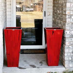 May include: Two tall, red, tapered planters stand on a concrete porch in front of a white door with a glass panel. The door has a sign that reads "No Soliciting".