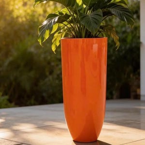 May include: A tall, vibrant orange planter filled with a lush green plant. The glossy, cylindrical planter stands on a tiled surface, with a blurred green background. The sunlight creates a warm, inviting atmosphere, highlighting the plant's leaves.