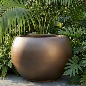 May include: A large, bronze-colored, spherical planter pot. The smooth, rounded pot sits on a stone surface, surrounded by lush green foliage. The planter is empty, ready for planting. The pot's color complements the natural greenery.