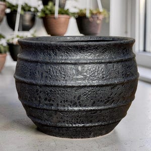 May include: A black, textured ceramic planter with a wide, rounded shape. The planter features horizontal bands with a detailed, speckled pattern. The pot is designed for indoor or outdoor use and is suitable for various plants.