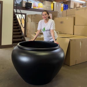 May include: A large, matte black, spherical planter with a wide, circular opening and a smooth, rounded body. The planter features a thick, reinforced rim and a deep, hollow interior, designed for holding plants or decorative arrangements.