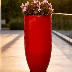 May include: A tall, cylindrical, glossy red planter with a smooth finish. The planter contains a cluster of small, light pink flowers with dark green leaves. A small, rectangular silver brand tag is attached to the upper rim.