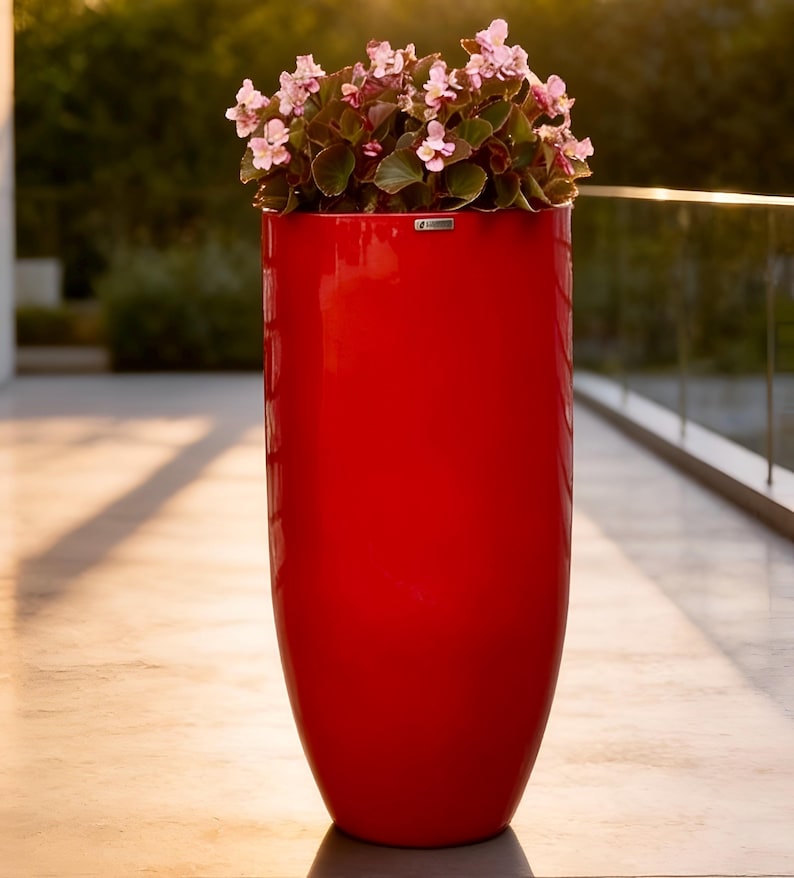 May include: A tall, cylindrical, glossy red planter with a smooth finish. The planter contains a cluster of small, light pink flowers with dark green leaves. A small, rectangular silver brand tag is attached to the upper rim.