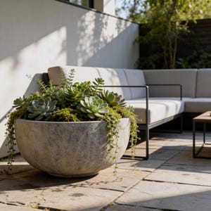 May include: A large, round, gray concrete planter filled with various green succulents and trailing plants. The planter sits on a stone patio, with a modern outdoor sofa in the background. The plants are a mix of textures and shades of green.