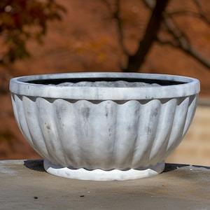 May include: A large, gray, fluted planter bowl with a wide, shallow design. The bowl has a scalloped rim and a rounded base. The exterior has a weathered appearance, with darker streaks adding depth. Ideal for outdoor or indoor use.