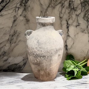 May include: A white ceramic vase with two handles, a rounded body, and a distressed finish. The vase is sitting on a white marble surface.
