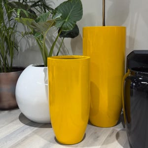 May include: Two tall, cylindrical, glossy yellow planters stand in the foreground. Behind them, a large, round, white planter holds a green plant with broad leaves. To the far left, a metallic bronze planter is visible, and to the right, a black planter.