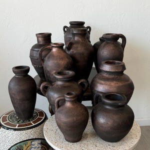 May include: A collection of nine brown ceramic vases of varying sizes and shapes. The vases are arranged on a white surface with a brown wooden table in the background.