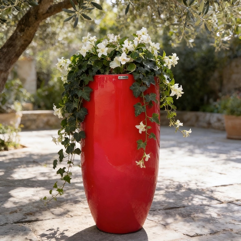 May include: A tall, cylindrical, glossy red planter with a smooth finish. The planter contains a lush arrangement of green ivy vines and clusters of small, white, five-petaled flowers that spill over the top edges of the container.