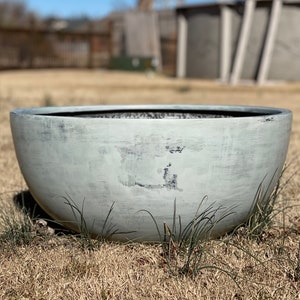 May include: A large, light gray, bowl-shaped planter with a distressed finish. The planter sits on dry grass, with a blurred background of a backyard and a swimming pool. The bowl is round and has a wide opening.