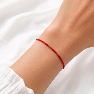 May include: A thin, vibrant red cord bracelet featuring a simple, minimalist design. The bracelet is constructed from a single, continuous strand of twisted red thread that wraps around the wrist, creating a clean and understated look for everyday wear.