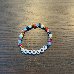 Peut inclure: Un bracelet en perles blanches avec des perles lettres noires qui épellent le mot "WAOLOM". Les perles sont entrecoupées de perles de cristal rouges, dorées et noires.