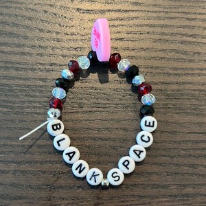 May include: A beaded bracelet with the word "BLANK SPACE" spelled out in white letter beads. The bracelet also features black, red, and clear beads, and a pink heart charm that says "BE MINE".