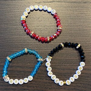 May include: Three beaded bracelets with white beads and gold lettering. The bracelets spell out the words "Screaming", "Crying", and "Perfect Storm".