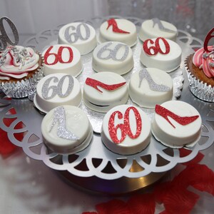May include: A silver cake stand with white and red decorated cupcakes and chocolate-covered cookies. The cupcakes and cookies are decorated with the number "60" and silver and red high heels.