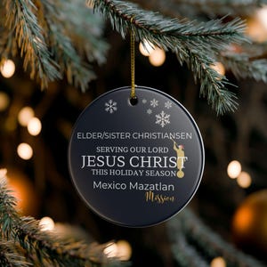 May include: A black round Christmas ornament with gold text and a gold string for hanging. The ornament has the text "ELDER/SISTER CHRISTIANSEN SERVING OUR LORD JESUS CHRIST THIS HOLIDAY SEASON Mexico Mazatlan Mission."