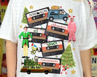 Könnte beinhalten: Weißes T-Shirt mit einem Weihnachtsfilm-Motiv. Das Design zeigt Kassetten mit den Titeln "Kevin - Allein zu Haus", "Die nassen Banditen", "Eine Weihnachtsgeschichte", "Der Weihnachtsmann", "Der Grinch" und "Weihnachten bei den Griswolds".