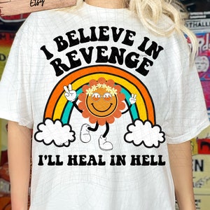 May include: White t-shirt with a rainbow and a smiling flower graphic. The text "I believe in revenge I'll heal in hell" is printed on the shirt.