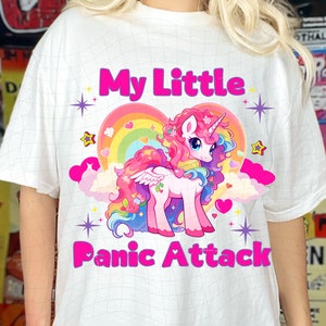 May include: White t-shirt featuring a pink unicorn graphic and the text "My Little Panic Attack". The unicorn is surrounded by a rainbow heart and stars.