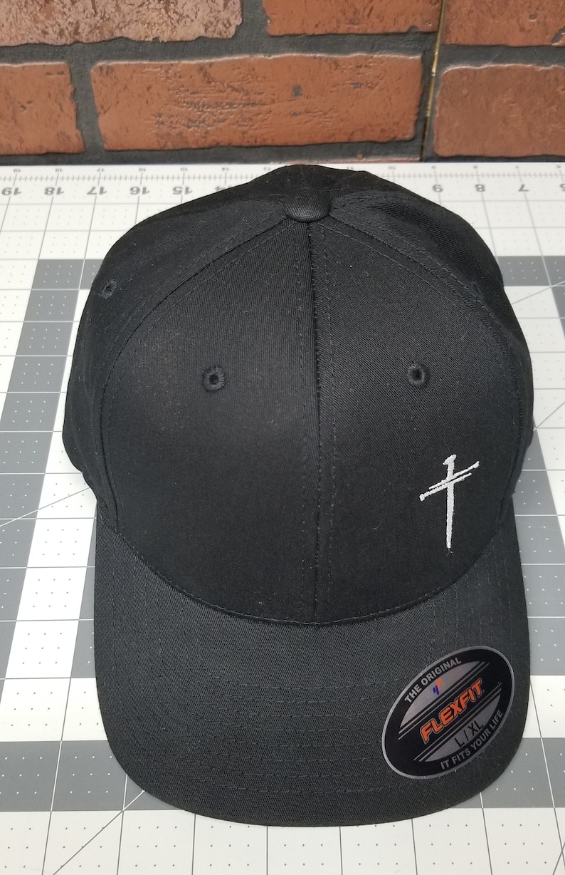 May include: Black baseball cap with a white cross embroidered on the front. The cap has a Flexfit logo on the side and says "The Original" and "It Fits Your Life".