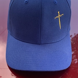 May include: A royal blue baseball cap with a gold embroidered cross on the front.