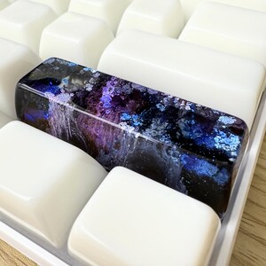 May include: A rectangular resin spacebar keycap featuring a deep cosmic design with swirling shades of dark blue, purple, and black, accented by shimmering light blue and silver flecks that resemble a nebula, set among plain white keyboard keys.
