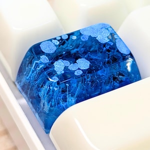 May include: A custom artisan keycap with a deep blue resin design, featuring lighter blue accents resembling a celestial or oceanic theme. The keycap is surrounded by white keycaps, creating a striking contrast.