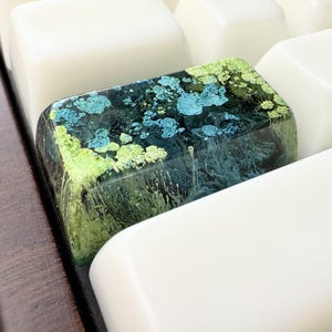 May include: A rectangular resin keycap featuring a translucent dark base with swirling patterns of vibrant lime green and bright turquoise ink. The keycap is positioned among solid white keys on a mechanical keyboard, displaying a smooth, glossy finish.