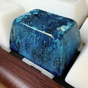 May include: A rectangular mechanical keyboard keycap featuring a deep blue resin base with swirling light blue and white ink patterns that resemble underwater currents or coral formations, set against a dark blue background with a glossy finish.